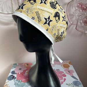 May include: A gold and black Vanderbilt Commodores scrub cap. The cap features the words "Vanderbilt Commodores" and "Anchor Down" along with star and hand symbols. The cap has a white trim and ties.