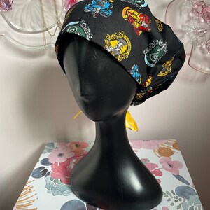 May include: Black surgical scrub cap featuring colorful crests from a fantasy series. The cap is displayed on a black mannequin head. The background includes decorative glass plates and a floral patterned surface.