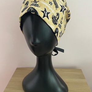 May include: A tan and black patterned scrub cap featuring the text "Commodores" and "Anchor Down" along with star and anchor graphics. The cap is displayed on a black mannequin head.
