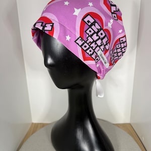 Könnte beinhalten: Eine OP-Haube mit rosa Hintergrund, roten Herzen, weißen Sternen und dem Text "Girls Run the World". Die Haube ist hinten mit weißen Bändern gebunden. Die Haube wird auf einem schwarzen Schaufensterkopf präsentiert.
