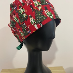 May include: A red patterned scrub cap with a Christmas theme. The cap features illustrations of nutcrackers and Christmas trees. A green ribbon is tied at the back. The cap is displayed on a black mannequin head.