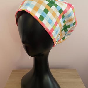 Op de afbeelding: Een kleurrijke scrub cap met patroon. De cap heeft een ruitjesontwerp in tinten oranje, groen, blauw en roze, met een roze rand. De cap wordt getoond op een zwarte mannequin hoofd.
