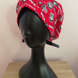May include: A red and white patterned scrub cap featuring the University of Georgia Bulldogs logo and text. The cap is displayed on a black mannequin head.