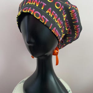 May include: Black scrub cap with the text "I AIN'T AFRAID OF NO GHOST" in a rainbow gradient. The cap has an orange ribbon tied at the back. The cap is displayed on a black mannequin head.