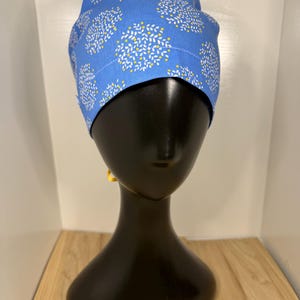 May include: A blue surgical scrub cap featuring a white and yellow floral design. The cap is made from a soft material and is designed to cover the head and hair. The cap is displayed on a black mannequin head.