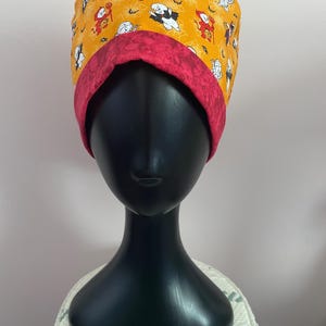 May include: A Halloween-themed scrub cap featuring a vibrant yellow background adorned with cartoon ghosts, devils, and spiderwebs. The cap has a contrasting deep red band. The cap is displayed on a black mannequin head.