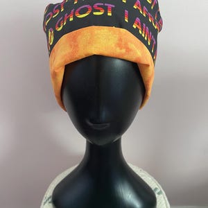 May include: Black surgical scrub cap with the text "I AIN'T A GHOST I AIN'T" in a rainbow gradient. The cap has an orange band and is displayed on a black mannequin head. The cap is made of fabric.