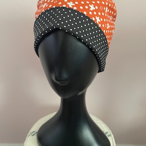 May include: A Halloween-themed turban-style hat. The top section is orange with white ghost shapes, and the bottom section is black with white polka dots. The hat is displayed on a black mannequin head.