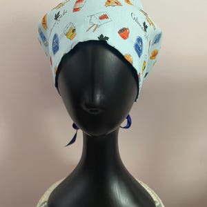 Puede incluir: Un gorro quirúrgico azul claro con un estampado de tazas de café, porciones de pizza y otros alimentos. El gorro también tiene las palabras "Gilmore Girls" impresas. El gorro se muestra en una cabeza de maniquí negra.