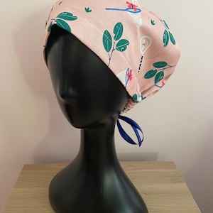 May include: A pink scrub cap with a repeating pattern of green leaves, white flowers, and blue accents. The cap is tied with a blue ribbon. The cap is displayed on a black mannequin head.