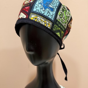 Könnte beinhalten: Eine gemusterte OP-Haube mit farbenfrohen Designs im Glasmalerei-Stil. Die Haube hat einen schwarzen Rand und Bindebänder. Der Stoff enthält Bilder eines Vogels, einer Schlange und das Wort "Slytherin".