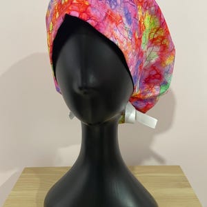 Pode incluir: Uma touca cirúrgica colorida com um padrão tie-dye vibrante em tons de rosa, vermelho, roxo e verde. A touca é exibida em uma cabeça de manequim preta e amarrada com uma fita branca.