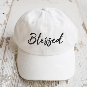 May include: A white baseball cap with a curved brim. The word "Blessed" is embroidered in black script across the front of the cap. The cap is made of a soft fabric and has a classic, casual style.