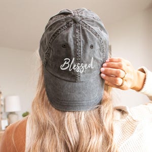 May include: A gray baseball cap with the word "Blessed" embroidered in white script. The cap is worn backward, and the brim is slightly curved. The hat appears to be made of a soft, worn material.