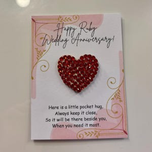 May include: A white card with a red jeweled heart and the text "Happy Ruby Wedding Anniversary!". The card also includes the text "Here is a little pocket hug..." in black font, with pink and gold decorative accents.