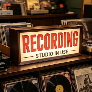 以下が含まれることがあります： 「RECORDING STUDIO IN USE」の赤い文字が書かれた長方形の木製サイン。サインは照らされており、レコードの棚に置かれています。サインはヴィンテージ感があります。