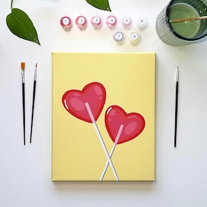 Op de afbeelding: Een paint-by-numbers kit met een canvas met twee rode hartvormige lolly's op een gele achtergrond. De kit bevat verfpotten, penselen en een glas water. De afbeelding staat tegen een witte achtergrond.