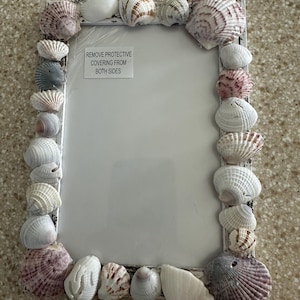 Könnte beinhalten: Weißer Bilderrahmen, der mit Muscheln verziert ist. Der Rahmen hat einen weißen Hintergrund und eine transparente Plastikabdeckung mit dem Text "REMOVE PROTECTIVE COVERING FROM BOTH SIDES".