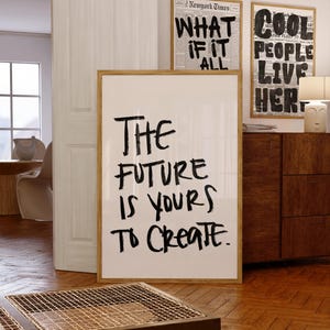 May include: A large rectangular framed wall art piece featuring the black handwritten text The Future Is Yours To Create on a white background. The frame is made of light-colored wood and stands upright on a wooden floor in a room setting.