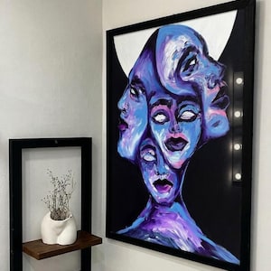 May include: Abstract painting featuring multiple faces in shades of blue and purple against a black background. The artwork is framed in black and mounted on a white wall. A small shelf with a white vase and dried plants is also visible.