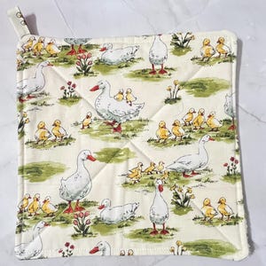 May include: A white quilted pot holder with a pattern of white ducks and yellow ducklings on a green background.