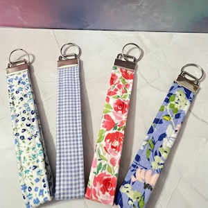 May include: Four keychains with different floral patterns. The keychains are made of fabric and have silver metal rings. The patterns include blue and white floral, blue and white gingham, pink and white floral, and blue and white floral with green leaves.
