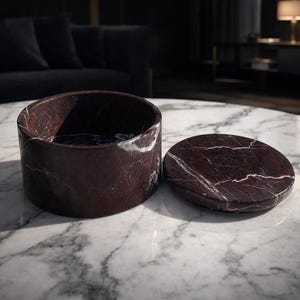 May include: A cylindrical, open-top container and a matching lid, both crafted from dark red marble with white veining. The items are displayed on a white marble table, suggesting a decorative or storage function.