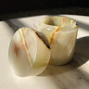 May include: A small, cylindrical jar with a lid, crafted from translucent, banded onyx. The jar is open, revealing the interior. The lid is placed beside the jar. The onyx features streaks of white, cream, brown, and orange.