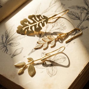 May include: Three gold-toned metal earrings shaped like botanical leaves. One earring features a wide, serrated oak-like leaf, while the other two are slender, elongated stems with smaller, delicate leaf attachments. Each piece has a curved hook.