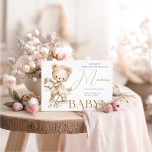 Peut inclure: Invitation de baby shower avec une illustration d'ours en peluche sur un cheval &agrave; bascule. Le texte dor&eacute; indique "Monica" et "oh BABY!" avec les d&eacute;tails de l'&eacute;v&eacute;nement. Pos&eacute;e sur une table en bois, orn&eacute;e de fleurs et de tissu rose.