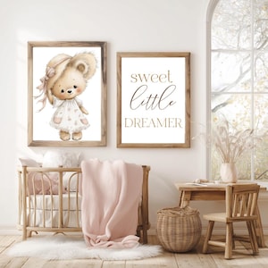 Peut inclure: Deux impressions encadr&eacute;es sur le th&egrave;me de la chambre d'enfant. L'une pr&eacute;sente une illustration &agrave; l'aquarelle d'un ours en peluche portant un chapeau et une robe. L'autre impression dit "sweet little dreamer" en lettres dor&eacute;es. Un berceau en bois, une couverture et un tapis sont au premier plan.