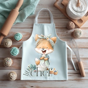 May include: A light blue apron with a cartoon fox illustration. The fox is wearing a headband and holding a whisk. The apron has the words "Little Chef" printed on it. Cupcakes and baking tools are arranged around the apron.