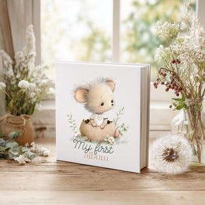 K&ouml;nnte beinhalten: Ein wei&szlig;es Fotoalbum mit einer Cartoon-Maus, die aus einem Ei schl&uuml;pft, umgeben von floralen Akzenten. Der Text "My first album" ist unter der Illustration aufgedruckt. Das Album wird auf einer Holzoberfl&auml;che mit Blumen und einem Fenster im Hintergrund pr&auml;sentiert.