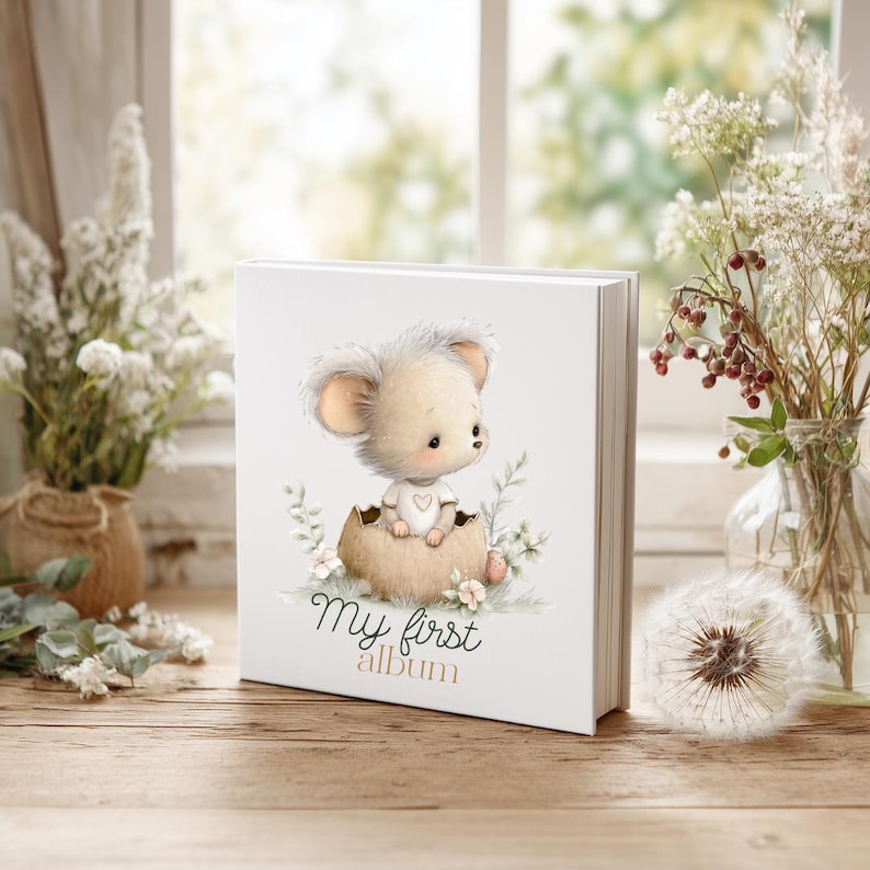 K&ouml;nnte beinhalten: Ein wei&szlig;es Fotoalbum mit einer Cartoon-Maus, die aus einem Ei schl&uuml;pft, umgeben von floralen Akzenten. Der Text "My first album" ist unter der Illustration aufgedruckt. Das Album wird auf einer Holzoberfl&auml;che mit Blumen und einem Fenster im Hintergrund pr&auml;sentiert.