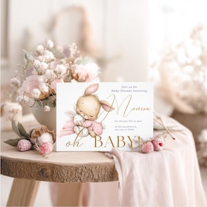 May include: A baby shower invitation featuring a watercolor illustration of a bunny hugging a smaller bunny. The invitation has gold text that reads "Monica" and "oh BABY!" on a white background. The invitation is on a wooden table with flowers.