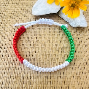 May include: A handmade bracelet with red, white, and green sections. The bracelet is made of woven cord and has an adjustable sliding knot closure. The bracelet is displayed on a woven surface with a yellow flower and white stones in the background.