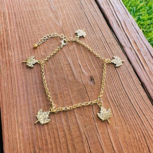 May include: Gold-colored anklet featuring five detailed maple leaf charms. The anklet is made of a delicate chain and has an adjustable length. The leaves are evenly spaced around the chain, creating a nature-inspired accessory.