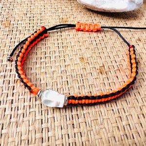 May include: A black and orange braided bracelet with a white skull charm. The bracelet is made of woven cord and features a sliding knot closure. The skull charm is made of a white material and is accented with silver-colored metal.