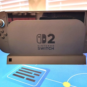 May include: A dark gray Nintendo Switch dock with the Nintendo Switch logo. The dock has a rectangular base and a slot to hold the console. The controllers are visible on either side of the screen. The background is a blue surface.