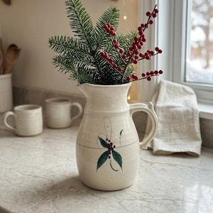 Peut inclure: Un pichet en céramique de couleur crème avec une finition mouchetée, orné d'un motif peint de feuilles vertes et de baies rouges sur le devant. Le pichet possède une anse incurvée et contient un arrangement décoratif de branches de pin.