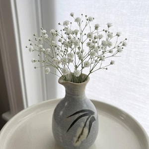May include: A small, grey ceramic vase with a black and white abstract design, holding a bouquet of white baby's breath flowers. The vase sits on a white, round tray, with a window in the background.