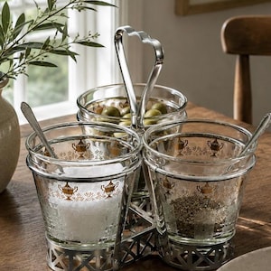May include: A three-part condiment server featuring clear glass bowls with gold urn motifs held in a silver-toned metal frame with a central heart-shaped handle. Two small silver spoons are included, with one bowl containing salt and another herbs.