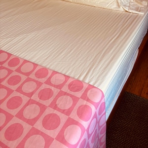May include: A bed with a cream-coloured striped sheet and two matching pillows. A pink and white patterned blanket with large circles and squares is draped over the foot of the bed. The bed is on a dark wood frame.