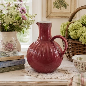 May include: A deep red ceramic pitcher with a rounded, ribbed body and a single curved handle. The pitcher features a glossy finish and a narrow neck with a small spout, standing as a central decorative object on a white surface.