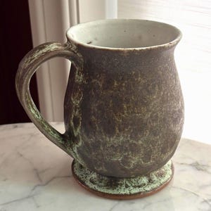 May include: A ceramic mug with a rounded body and a curved handle. The exterior features a textured, mottled brown and dark green glaze, while the interior is finished in a smooth, off-white glaze. The mug sits on a circular, light green base.