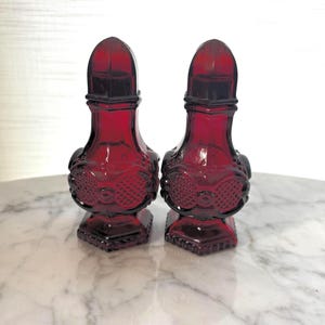 May include: A pair of deep red glass salt and pepper shakers featuring a moulded, textured pattern on the rounded bodies, a narrow neck, and a dome-shaped top. The shakers have a hexagonal base and a translucent finish that catches the light.