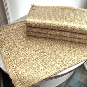 Peut inclure: Sets de sets de table beiges tissés avec des bords à franges. Un set de table est posé à plat, tandis qu'une pile de trois autres repose dessus. Les sets de table ont un tissage texturé et une couleur neutre.