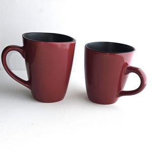 Peut inclure: Une tasse en céramique blanche avec une anse noire et le texte "But First, Coffee" en gras noir. La tasse est posée sur un fond flou, suggérant un cadre chaleureux.