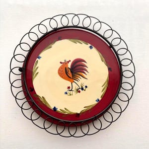 May include: A decorative plate with a rooster design. The plate has a cream-coloured centre with a rooster illustration, surrounded by a red border and a black metal frame. The rooster is orange and red, with blue and green accents.