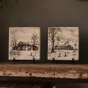 May include: Two square ceramic tiles featuring black and white ink-style landscape illustrations of rustic barns and rural fencing. Each tile is displayed upright on a small black metal stand, showcasing detailed sketches of trees and open fields.